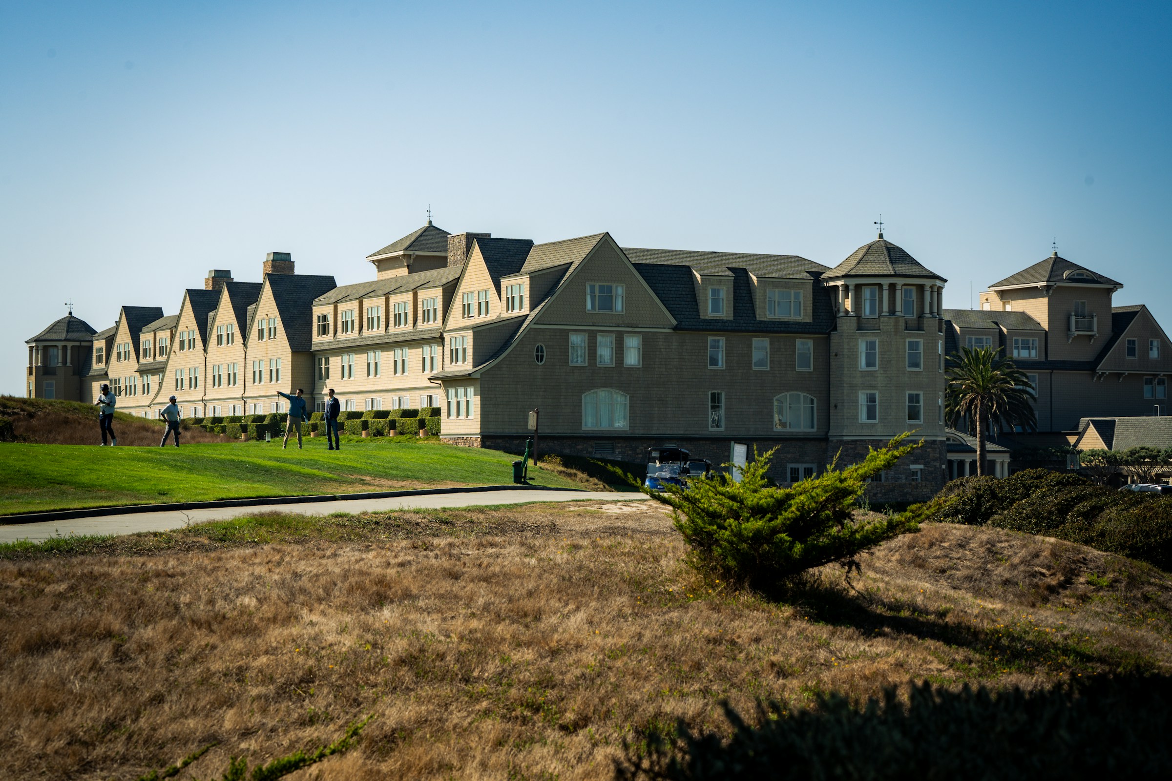 A large shingled resort hotel on a grass bluff above the California coast, with a few people standing on the lawn in front of it