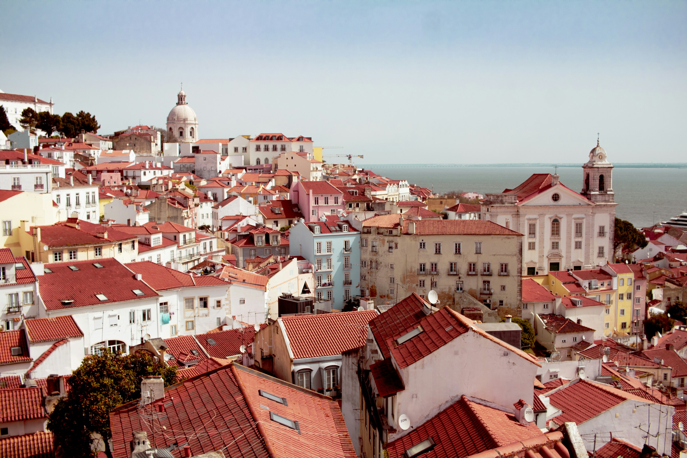 Pale houses and tiled roofs stacked on the hillside above the water in Lisbon, Portugal