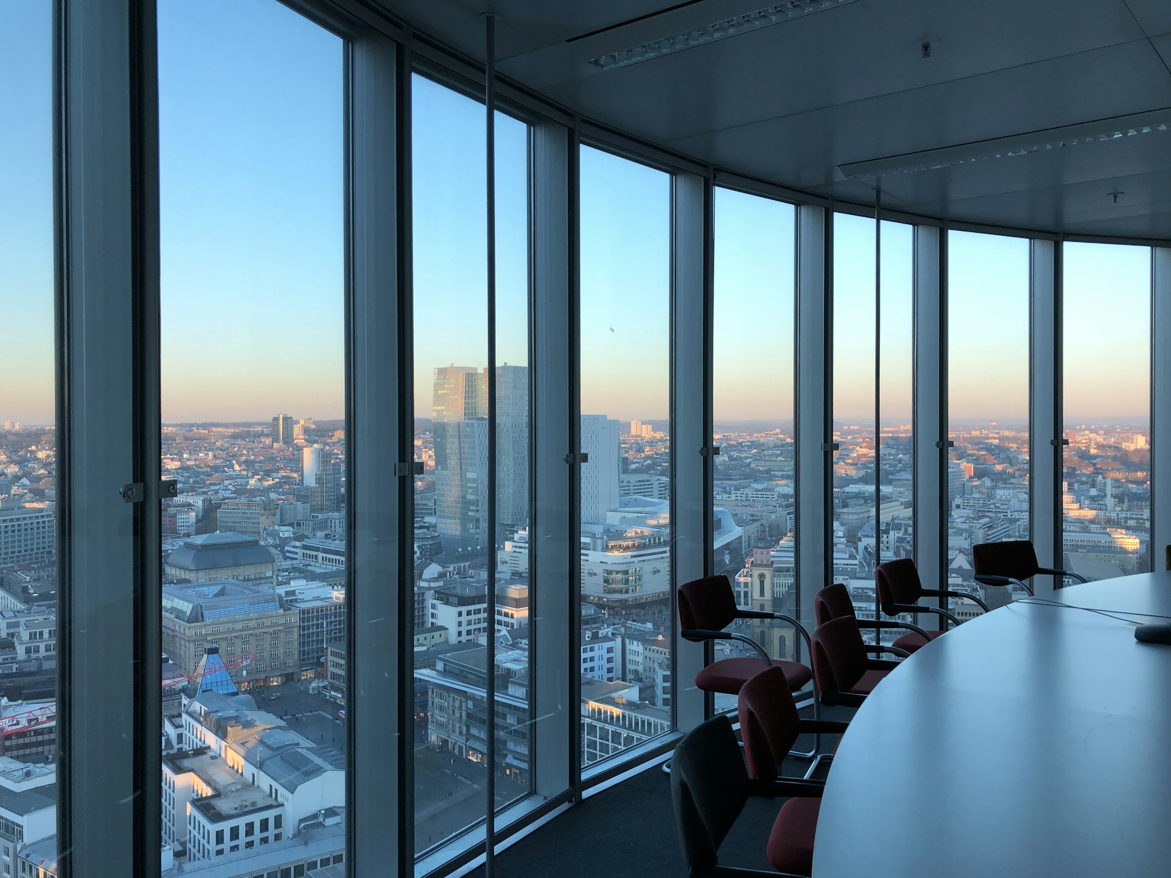 A corner boardroom high in a tower, chairs along a white table and glass walls looking out over a city at dusk