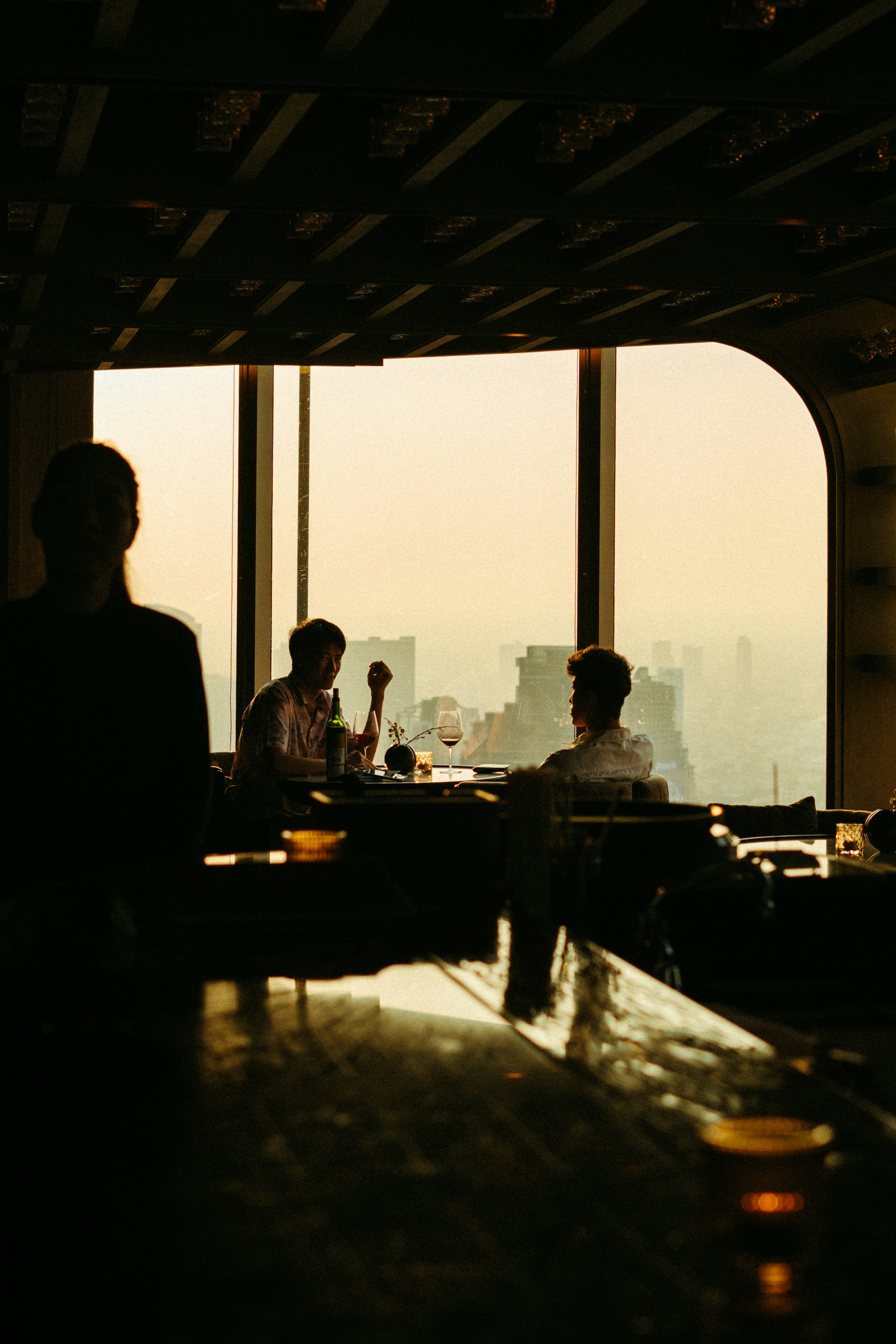 Two people talking over a table by a tall window in a high-floor bar, a hazy city skyline behind them at golden hour