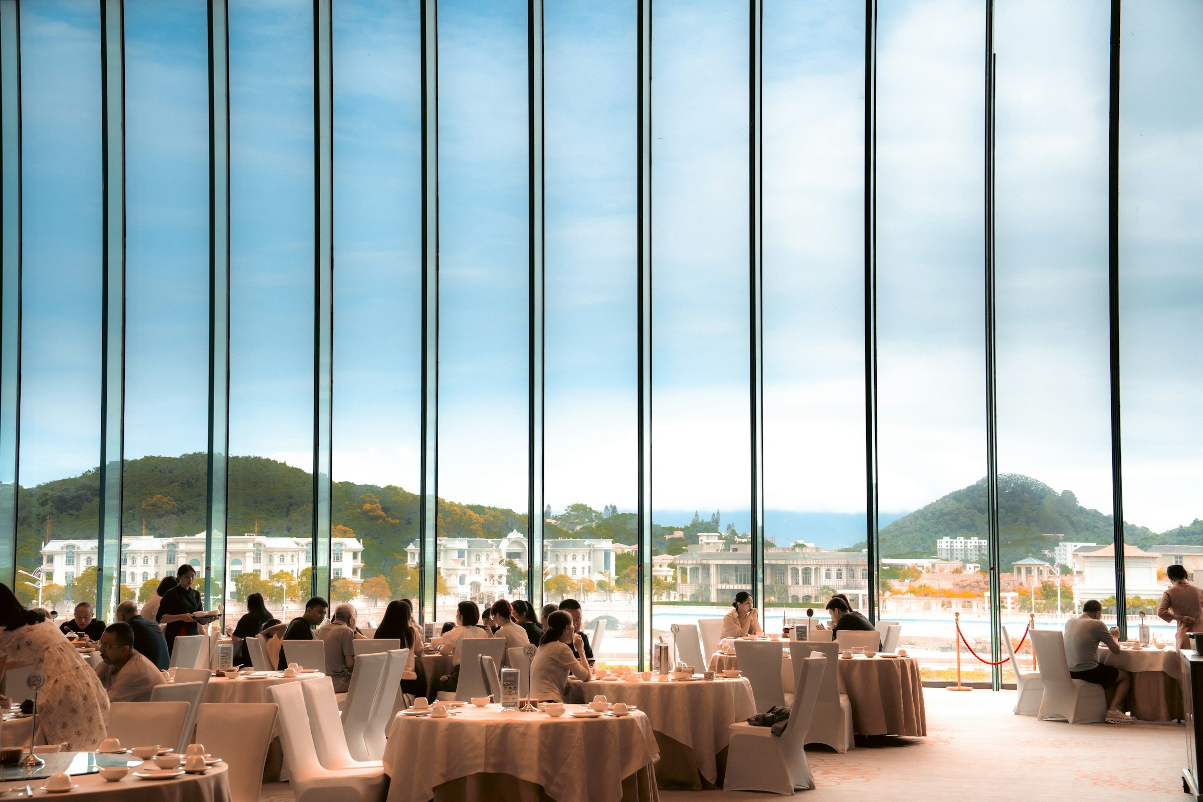 Diners at round tables in a restaurant with floor-to-ceiling windows looking out over a landscape